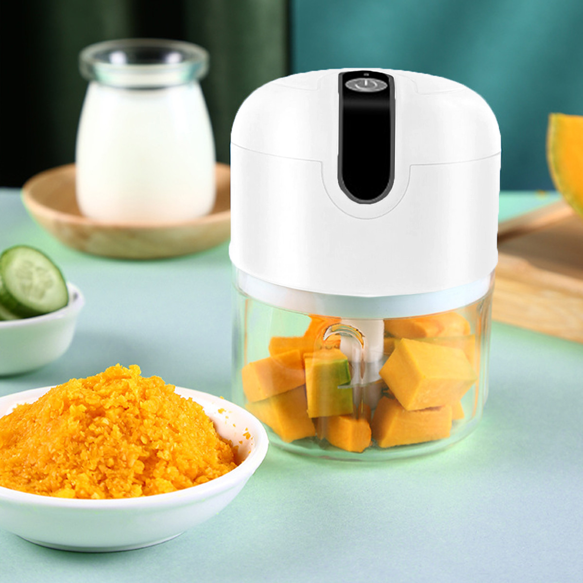 Ein weißer Foodprozessor mit Kürbis, eine Schüssel mit orangenem Essen und Gurkenscheiben, mit einem Milchglas im Hintergrund.