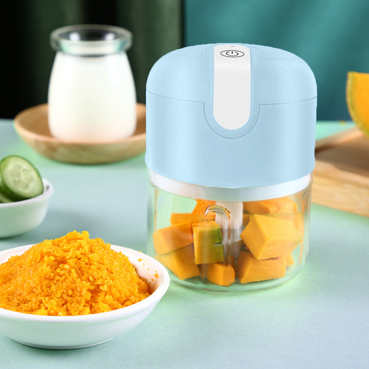 Ein Food Processor mit gehacktem Kürbis, Kürreepüree in einer Schüssel, Gurkenscheiben und ein Glas Milch auf einem Holztablett.