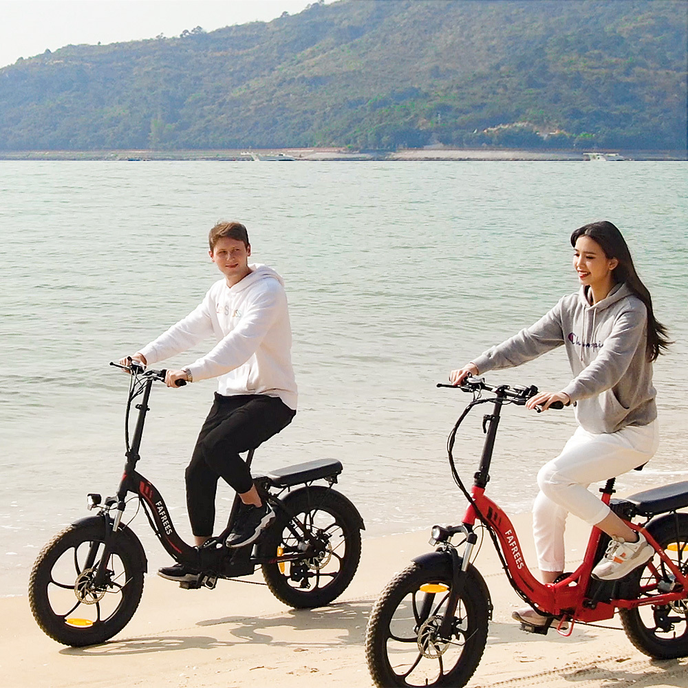Zwei Personen fahren mit Elektrofahrrädern am Strand. Ein Fahrrad ist rot, das andere schwarz. Im Hintergrund sind ein Berg und das Meer.