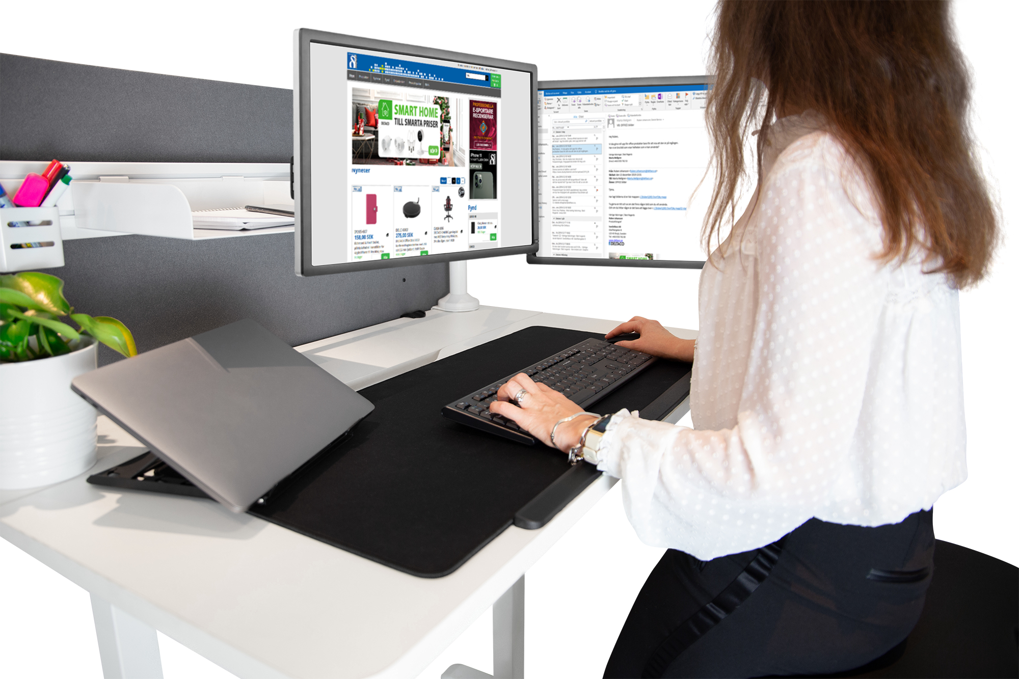 Eine Frau sitzt an einem Schreibtisch mit zwei Monitoren und tippt auf einer Tastatur. Ein Laptop, eine Topfpflanze und Schreibtischzubehör sind ebenfalls vorhanden.