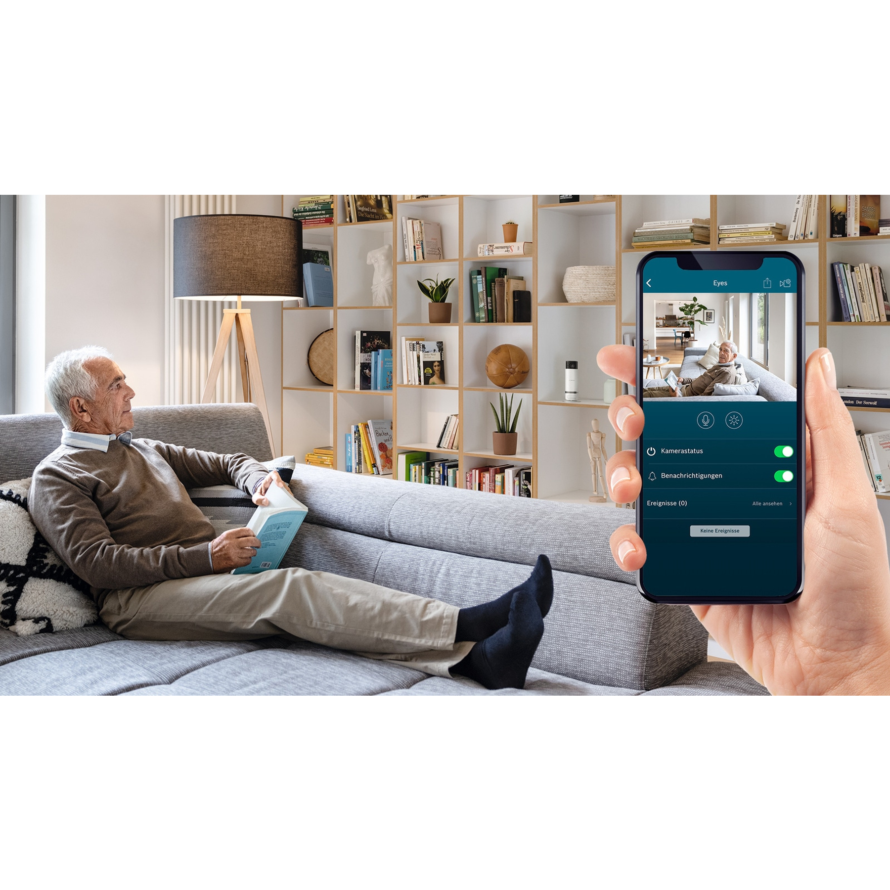 Ein älterer Mann liest auf einem Sofa. Ein Telefon zeigt ihn, gesehen von einer Kamera. Bücherregale sind im Hintergrund.
