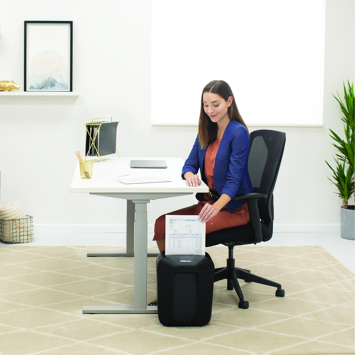 Kobieta siedzi przy biurku i niszczy dokument. Ma na sobie niebieską marynarkę. Czarna niszczarka stoi u jej stóp. Otoczenie biurowe.