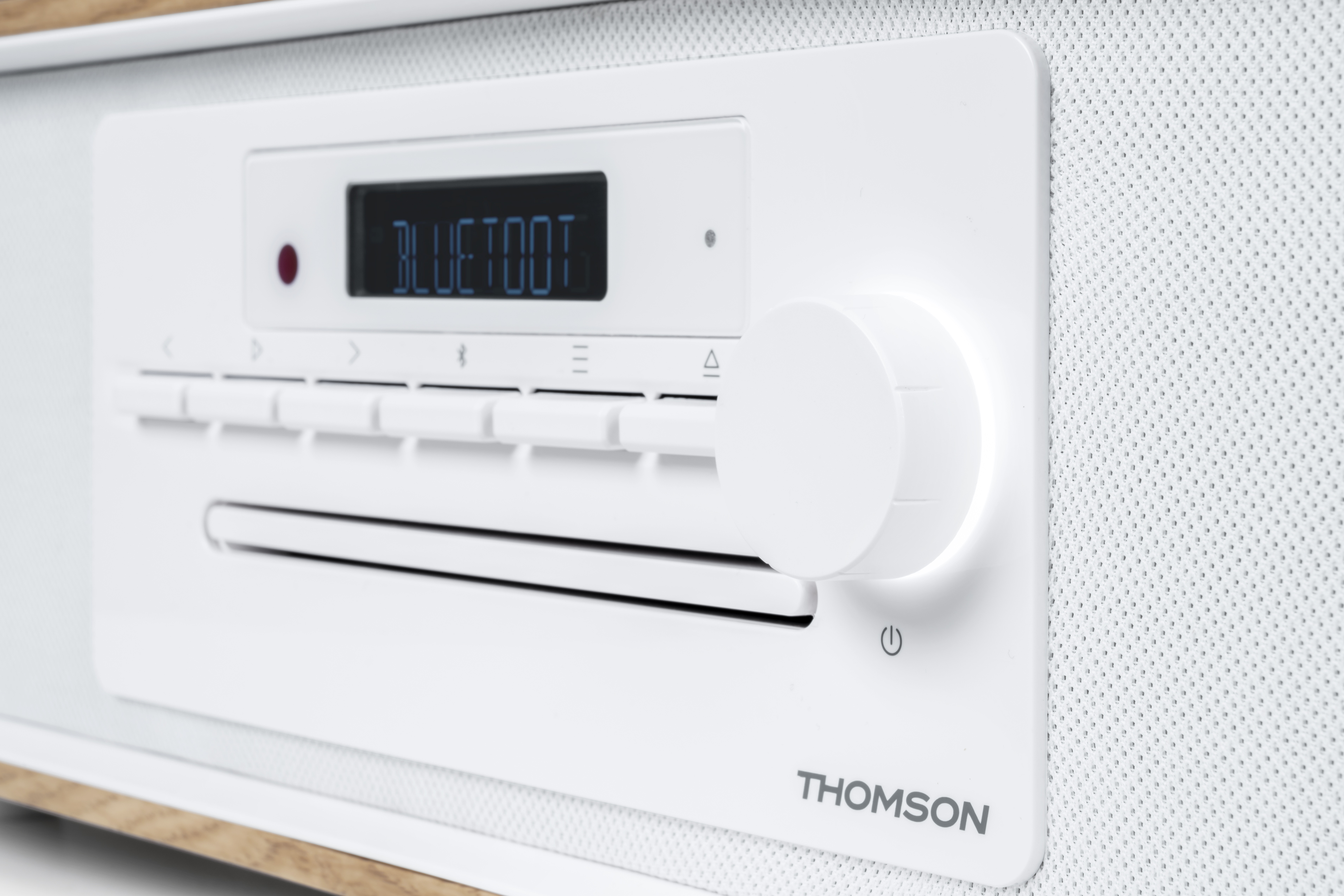 Białe stereo z wyświetlaczem 'BLUETOOT', przyciskami i pokrętłem. Tekst brzmi 'THOMSON'. Tło to biała tkanina.