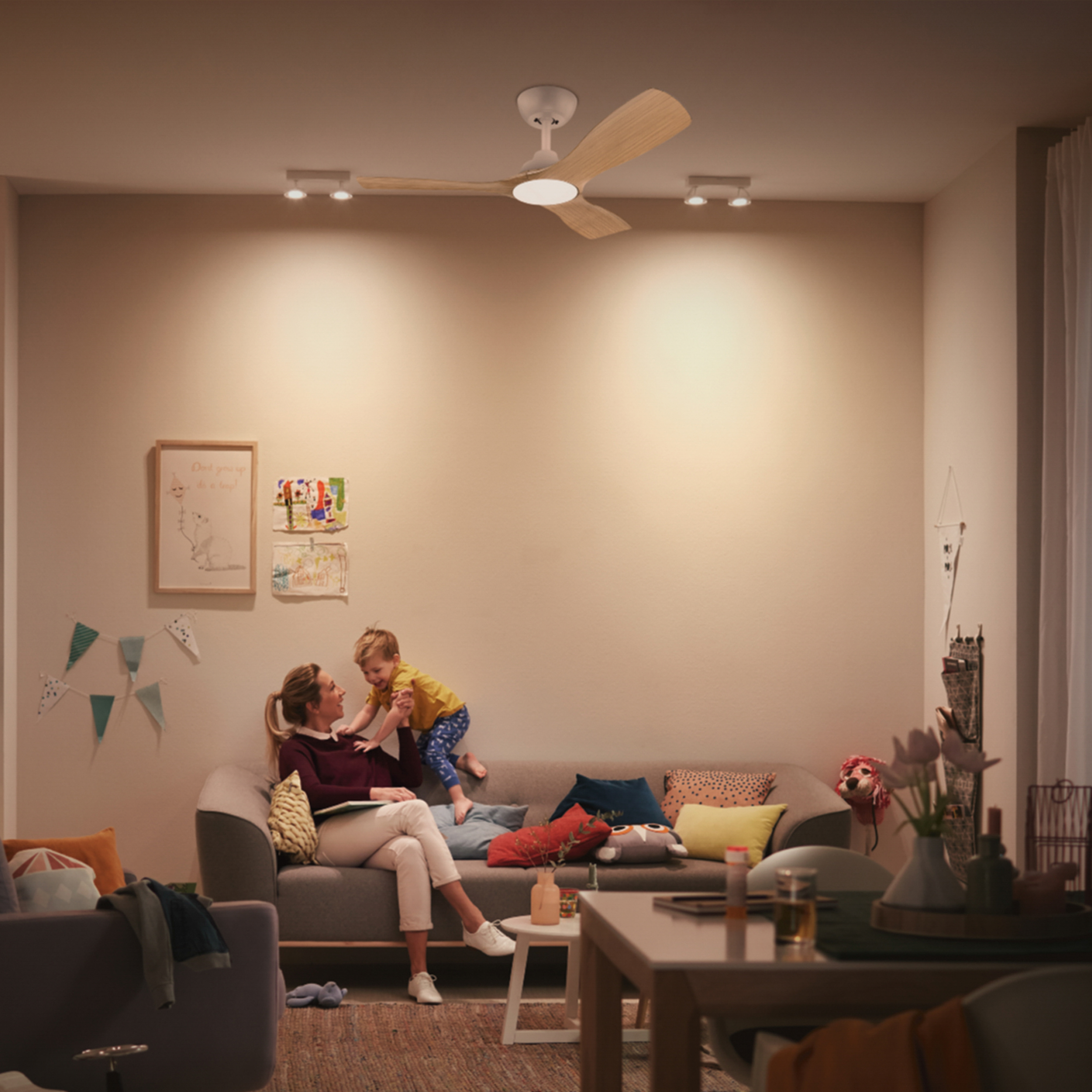 Una mujer y un niño están en un sofá en una habitación iluminada. Se ven un ventilador de techo y dos focos.