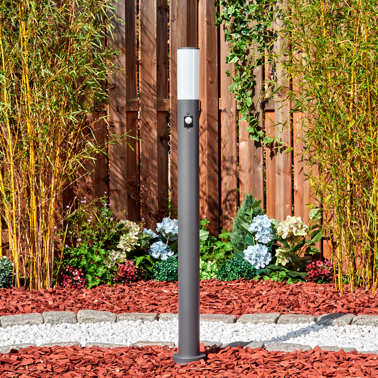 Eine graue Außenlampe steht in einem Gartenbeet mit roter Mulch und Blumen, mit einem Holzzaun und Bambus im Hintergrund.