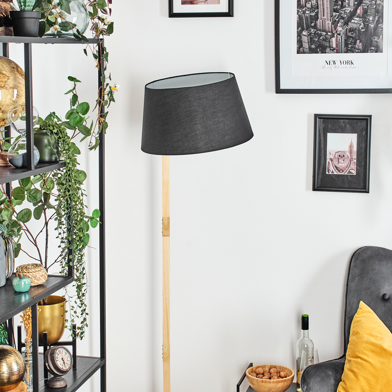 Ein Raum mit einer schwarzen Lampe, einem Regal mit Pflanzen, gerahmten Bildern an der Wand und einem Stuhl mit gelbem Kissen.