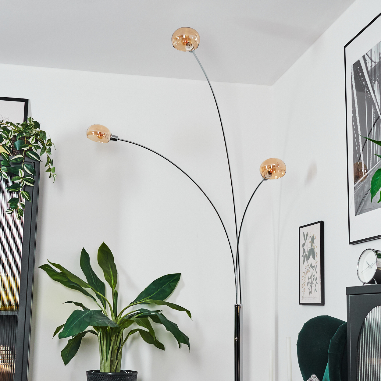 Eine moderne Stehlampe mit drei bernsteinfarbenen Kugeln. Sie steht in einem Raum mit Pflanzen, Kunstwerken und einer Uhr. Die Wand ist weiß.