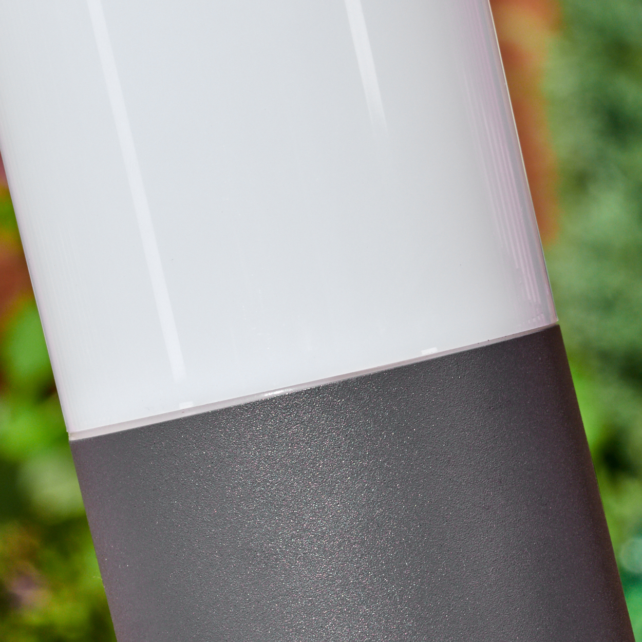 Nahaufnahme einer zylindrischen Lampe. Die obere Hälfte ist weiß, die untere Hälfte grau. Grünes Laub ist im Hintergrund unscharf.