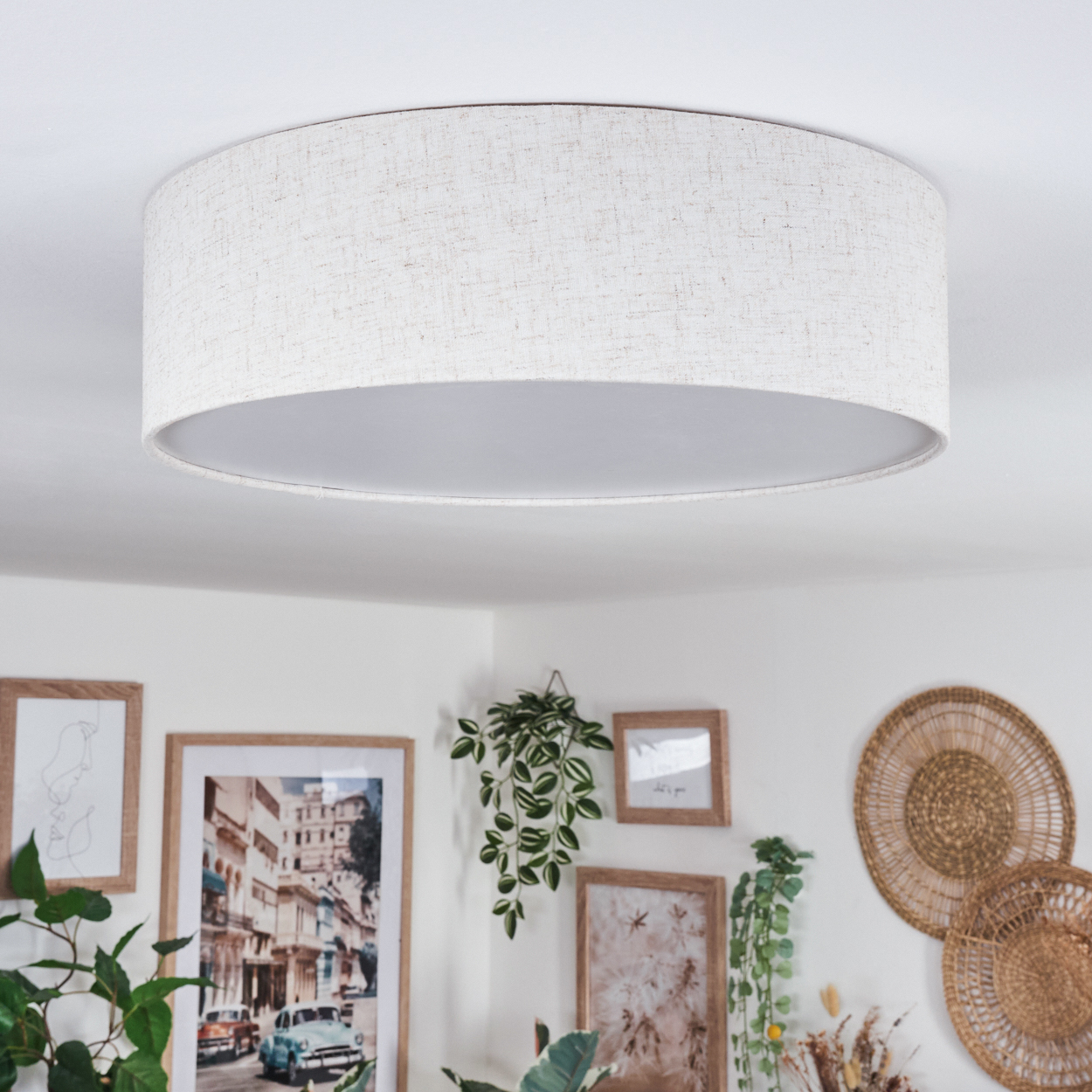 Eine große, beige, kreisförmige Deckenlampe ist zentriert. Darunter schmücken gerahmte Bilder und Hängepflanzen eine weiße Wand. Geflochtene Körbe sind vorhanden.