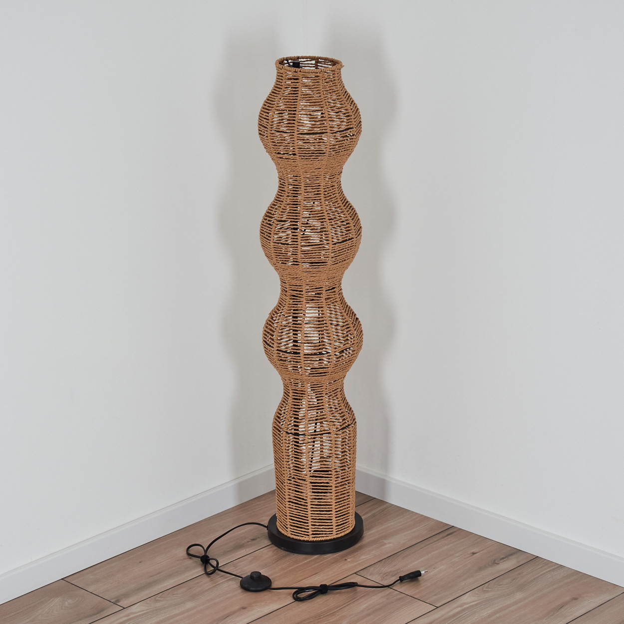 Eine gewebte, braune Stehlampe mit schwarzem Sockel steht in einer Ecke. Die Wände sind weiß und der Boden ist Holz.