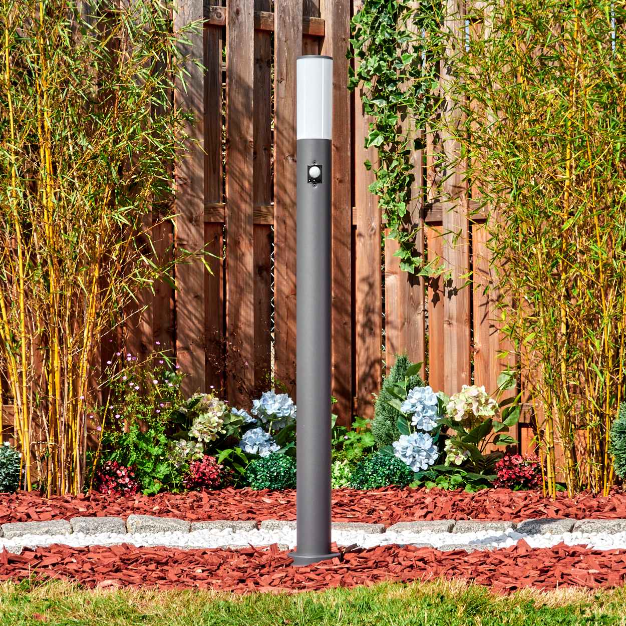 Eine graue Außenlampe steht in einem Garten. Bambus, Blumen und ein Holzzaun sind im Hintergrund. Roter Mulch und grünes Gras sind sichtbar.