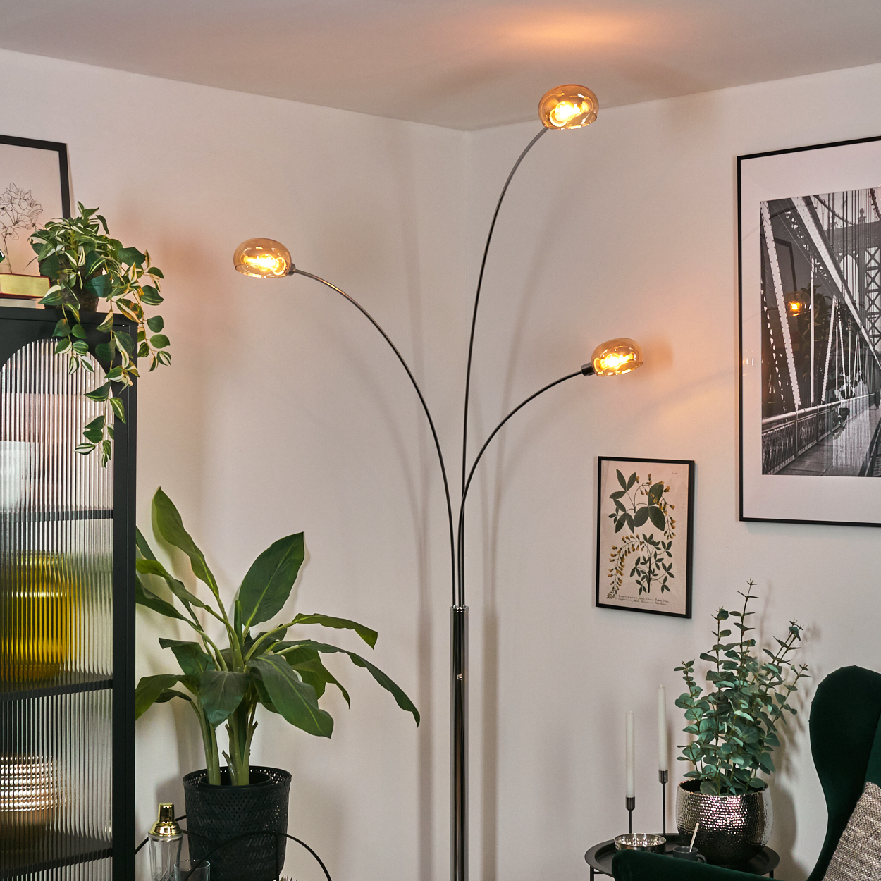 Ein modernes Wohnzimmer mit schwarzer Lampe, Pflanzen und gerahmter Kunst. Die Lampe hat drei Lichter. Pflanzen in schwarzen Töpfen.