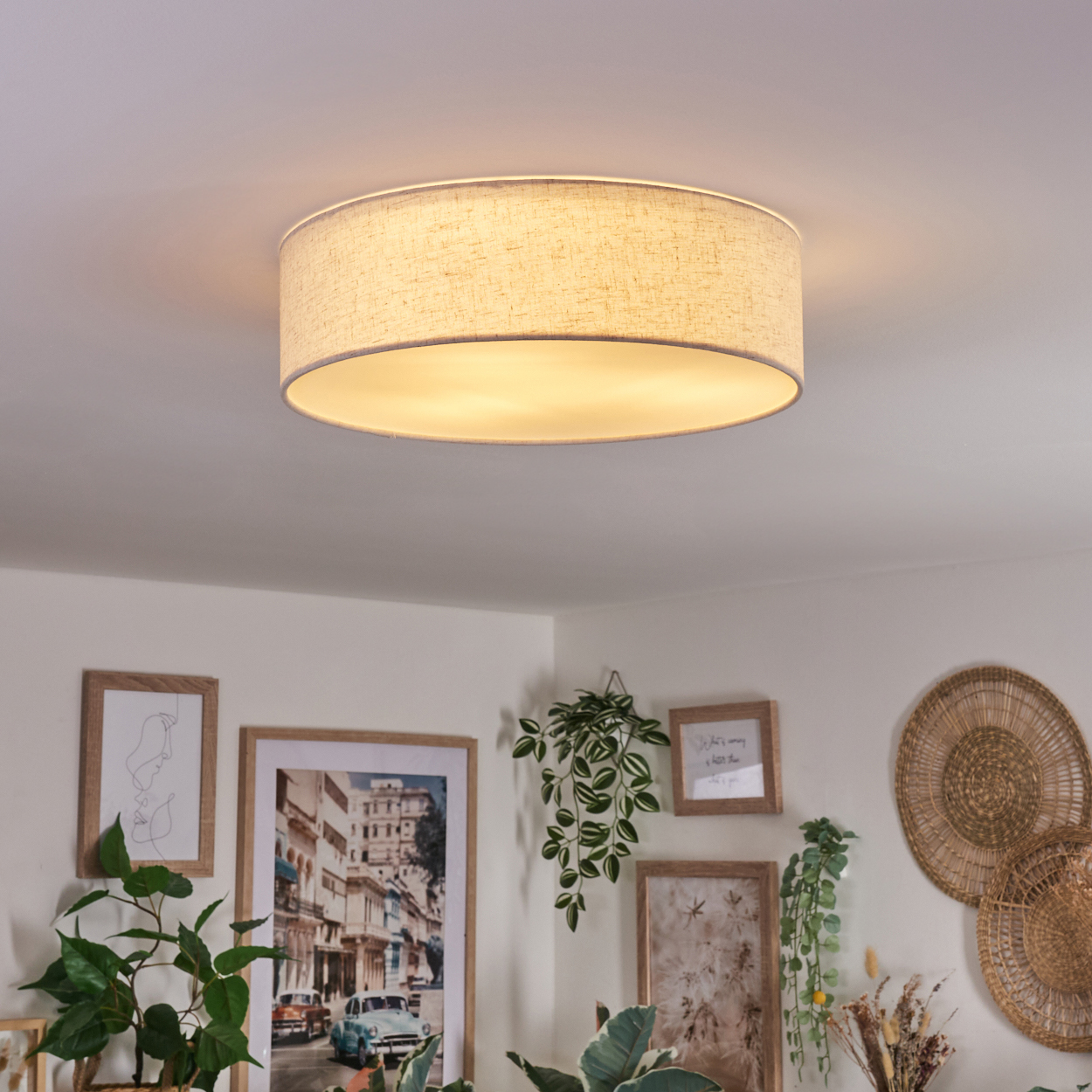 Eine runde Deckenlampe mit Leinenschirm leuchtet. Im Hintergrund sind Bilder und Pflanzen.
