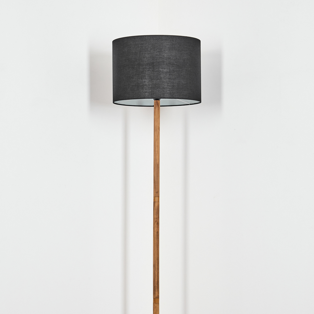 Eine Stehlampe mit schwarzem Lampenschirm und Holzstange steht vor einer weißen Wand. Schatten werden auf die Wand geworfen.