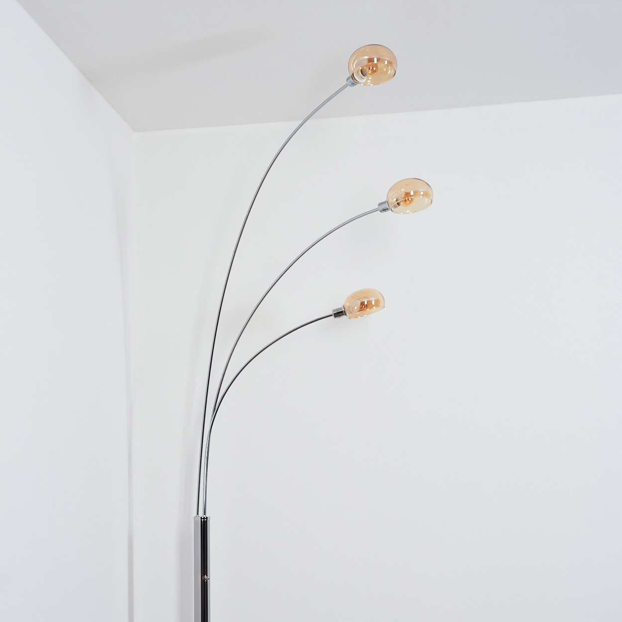 Eine moderne Stehlampe mit drei gebogenen Armen, die jeweils eine runde, bernsteinfarbene Leuchte halten. Der Hintergrund ist eine weiße Wand.