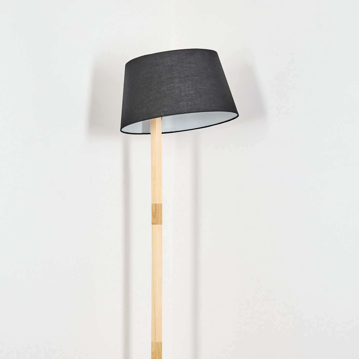 Eine Stehlampe mit schwarzem Lampenschirm und Holzfuß steht vor einer weißen Wand. Der Lampenschirm ist geneigt.