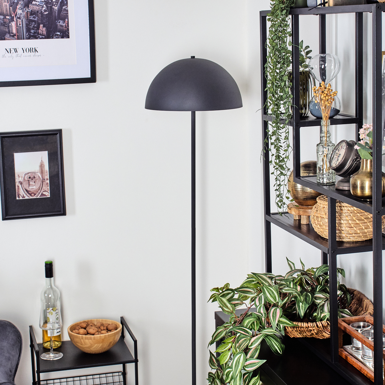 Ein Raum mit einer schwarzen Stehlampe, einem Regal mit Dekorationen und einem kleinen Tisch mit einer Schüssel Nüssen und einer Weinflasche.