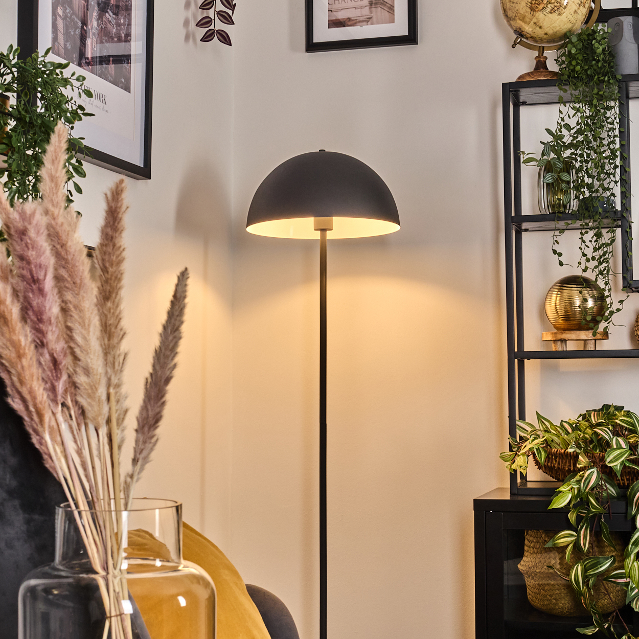 Eine dunkelgraue Stehlampe beleuchtet eine Ecke. Pampasgras in einer Vase und ein schwarzes Regal mit Pflanzen und einem Globus sind sichtbar.