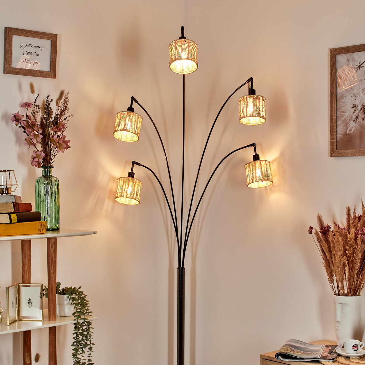 Eine Stehlampe mit fünf gewebten Schirmen. Das Zimmer hat eine weiße Wand, ein Regal mit Büchern und eine Vase mit Blumen.