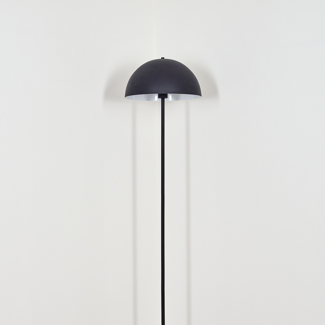 Eine schwarze Stehlampe mit halbkugelförmigem Schirm steht vor einer weißen Wand. Die Lampe hat einen geraden schwarzen Stab.