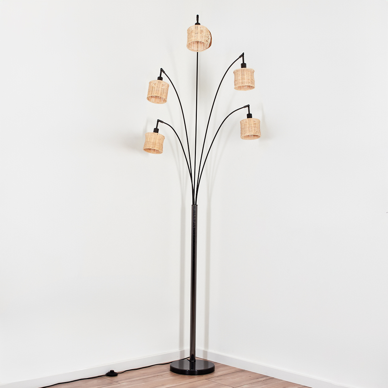 Eine moderne Stehlampe mit fünf gewebten Lampenschirmen. Die Lampe hat ein schwarzes Metallgestell und steht in einer Ecke an einer weißen Wand.