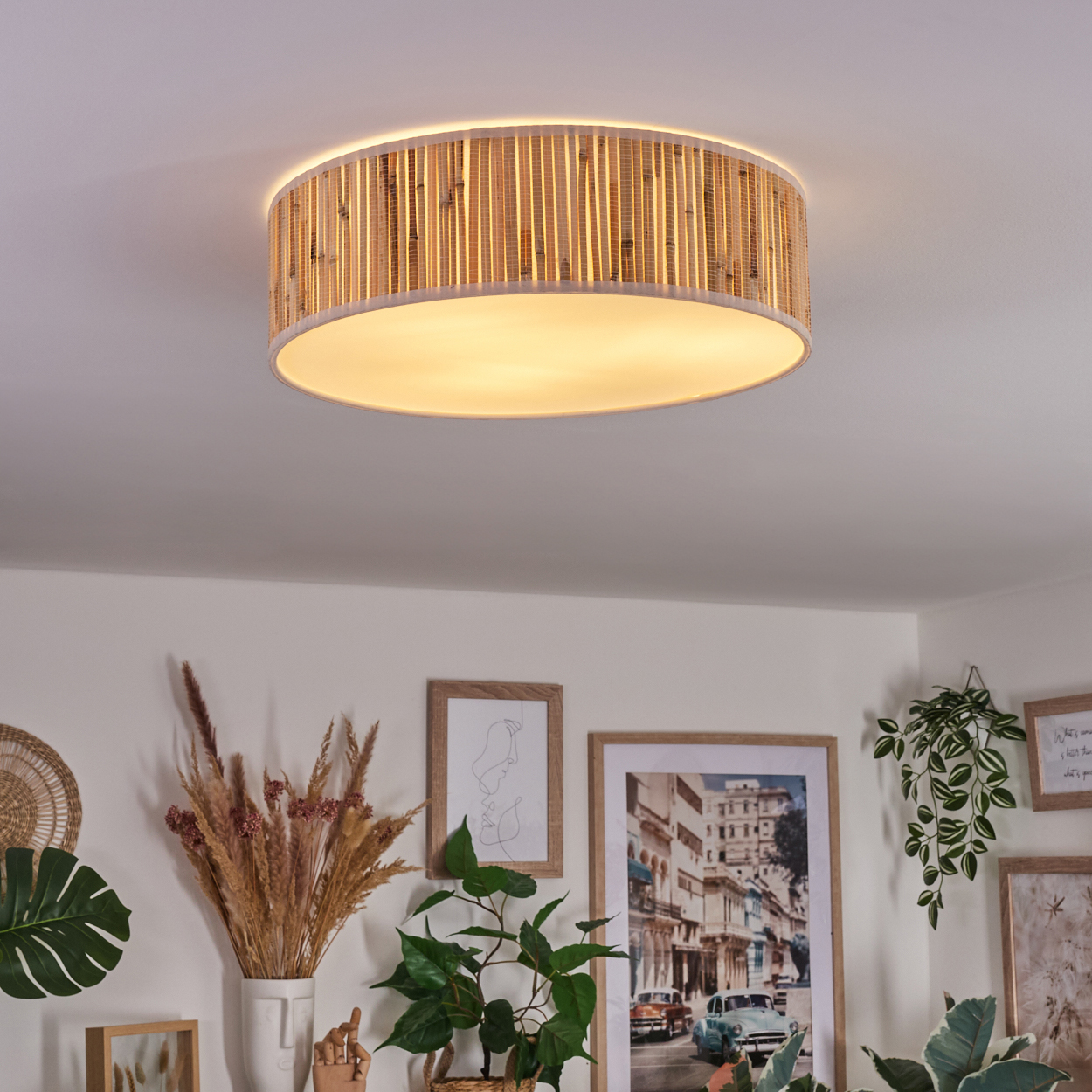 Eine Deckenlampe mit Bambusschatten leuchtet. Darunter dekorieren verschiedene gerahmte Bilder und Pflanzen die weiße Wand.