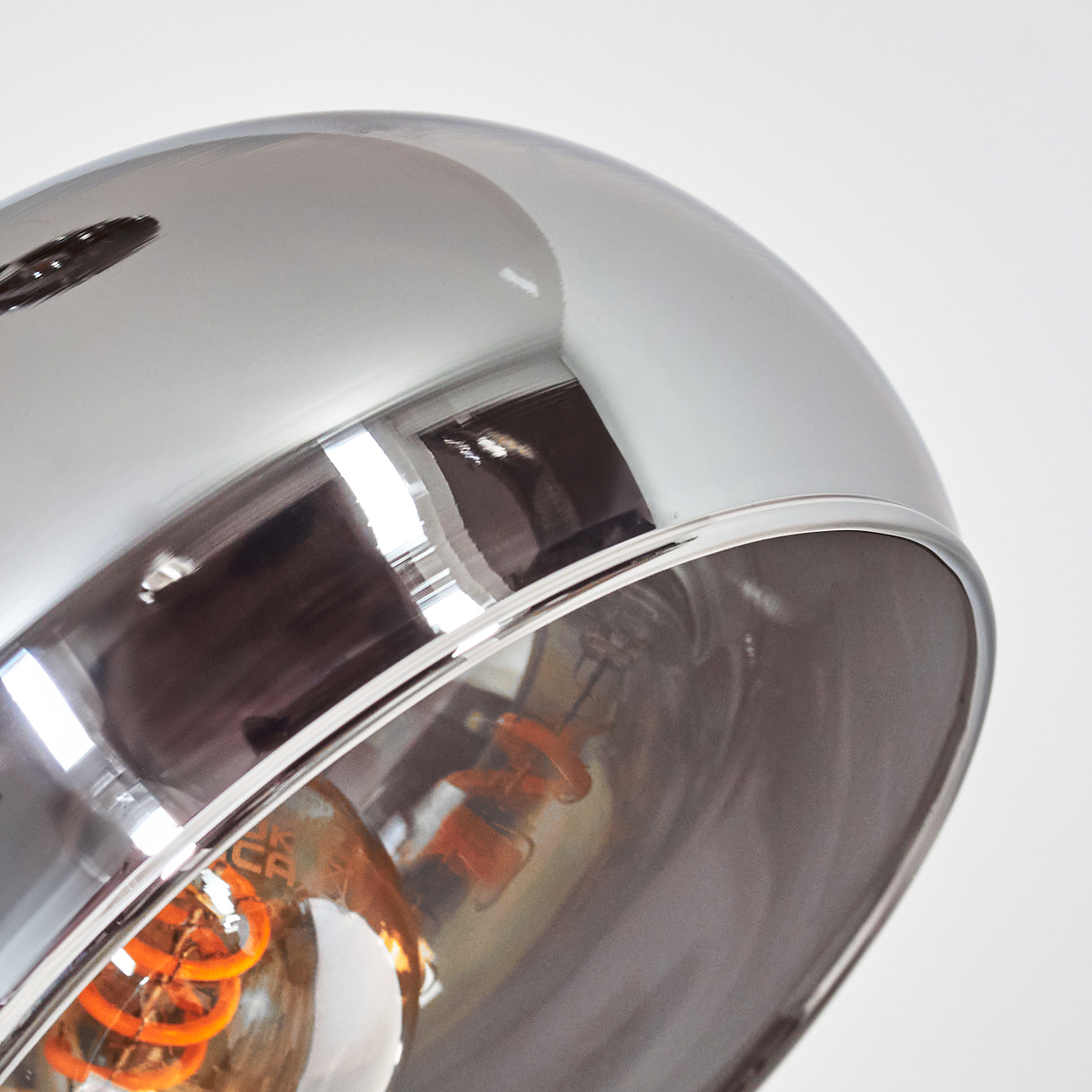 Nahaufnahme einer glänzenden Chromlampe. Im Inneren ist eine Glühbirne mit orangefarbenem Wendel sichtbar, die Licht reflektiert.