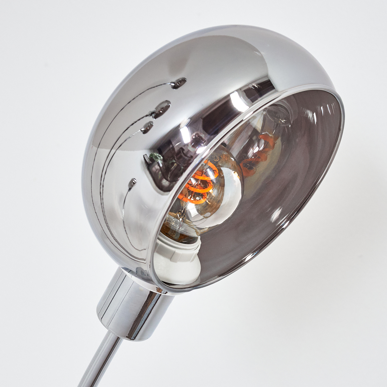 Chrom-Schreibtischlampe mit freiliegender Glühbirne mit orangefarbenem Glühfaden. Die Lampe reflektiert das Licht, vor weißem Hintergrund.