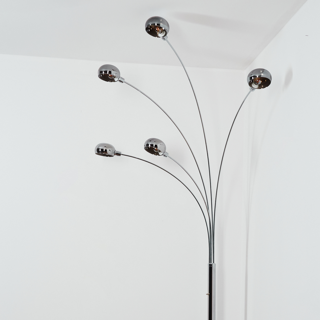 Eine verchromte Stehlampe mit fünf Lichtern. Jedes Licht ist eine Chromkugel. Die Lampe steht vor einer weißen Wand.