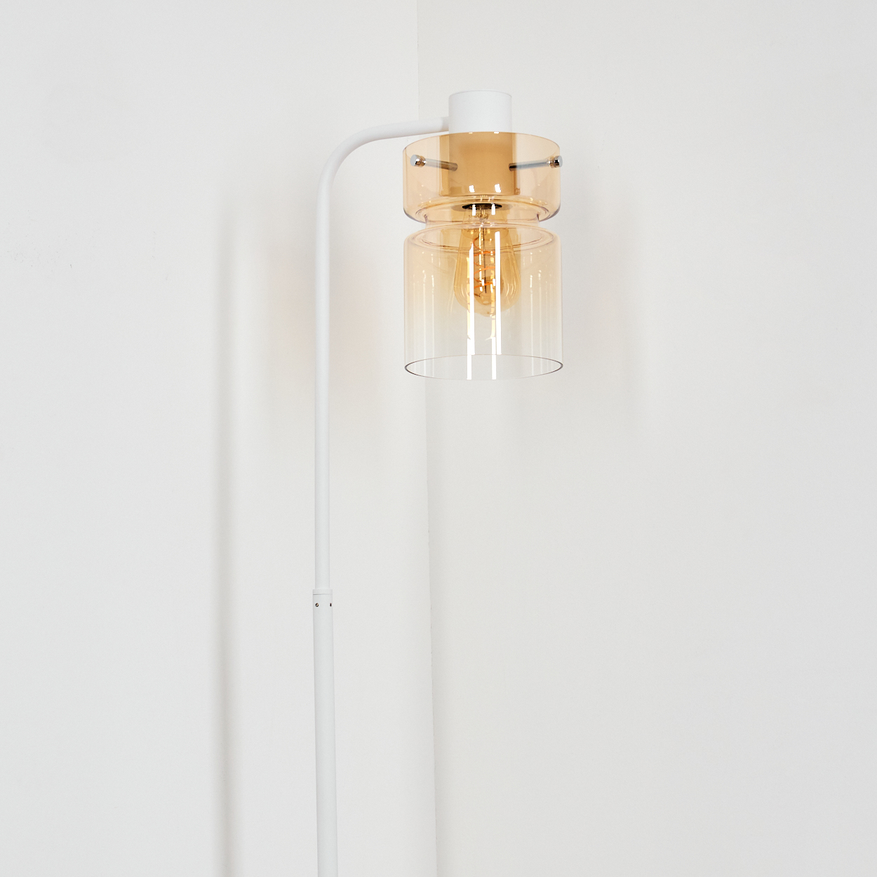 Eine weiße Stehlampe mit gebogenem Arm trägt einen Glasschirm mit gelber Glühbirne vor weißer Wand.