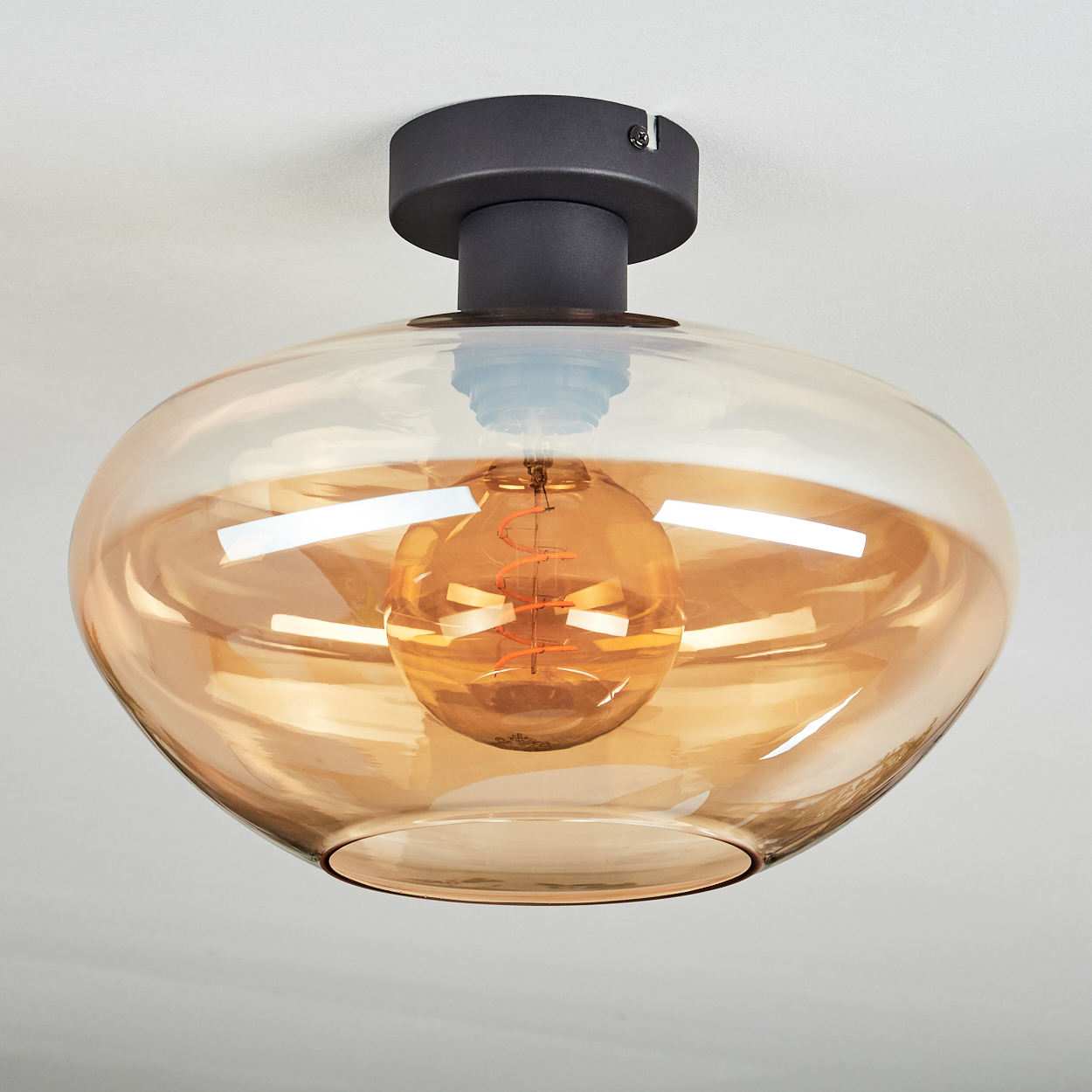 Eine moderne Deckenleuchte. Sie hat einen abgerundeten, bernsteinfarbenen Glasschirm und einen dunklen Metallsockel. Der Hintergrund ist eine weiße Decke.