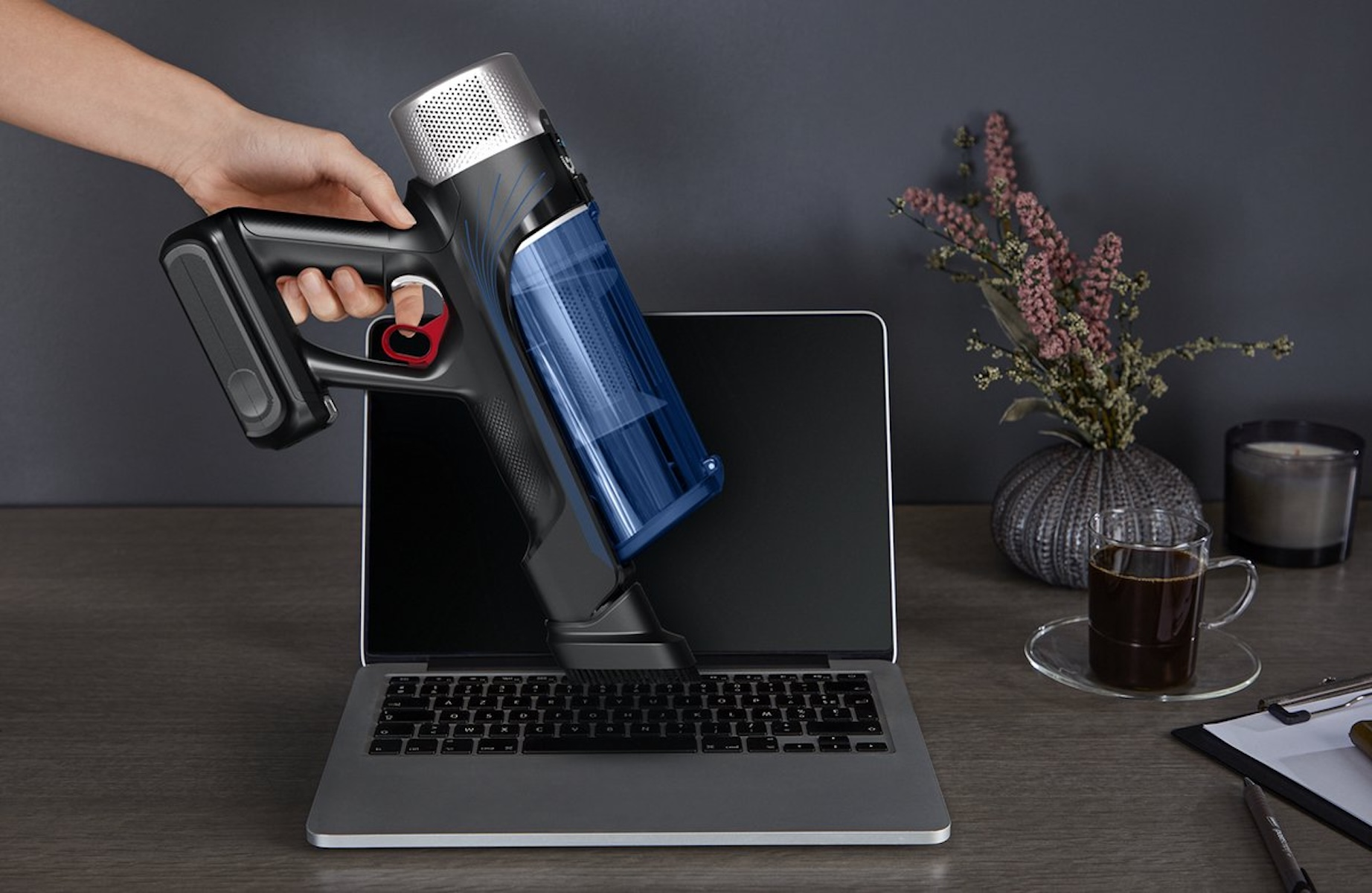 Een hand houdt een stofzuiger boven een laptop. Er staat een vaas met bloemen, een kopje koffie en een kaars op het bureau.