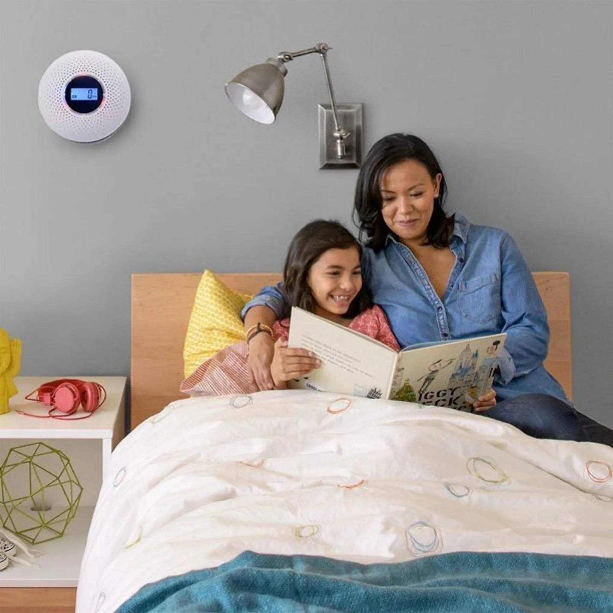 Eine Frau und ein Mädchen lesen ein Buch auf einem Bett. Ein Rauchmelder und eine Lampe sind an der Wand. Kopfhörer und eine Skulptur stehen auf einem Tisch.