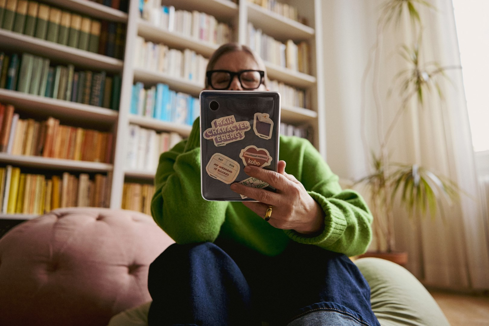 Osoba w okularach i zielonym swetrze trzyma telefon z naklejkami. W tle regały z książkami, różowa poduszka i okno z zasłonami.