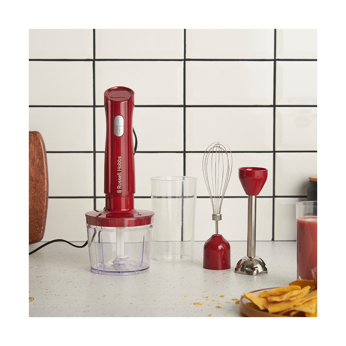 Ein roter Russell Hobbs Stabmixer mit Zubehör steht auf einer Arbeitsplatte. Ein Glas, ein Schneebesen und ein Trinkglas sind sichtbar.