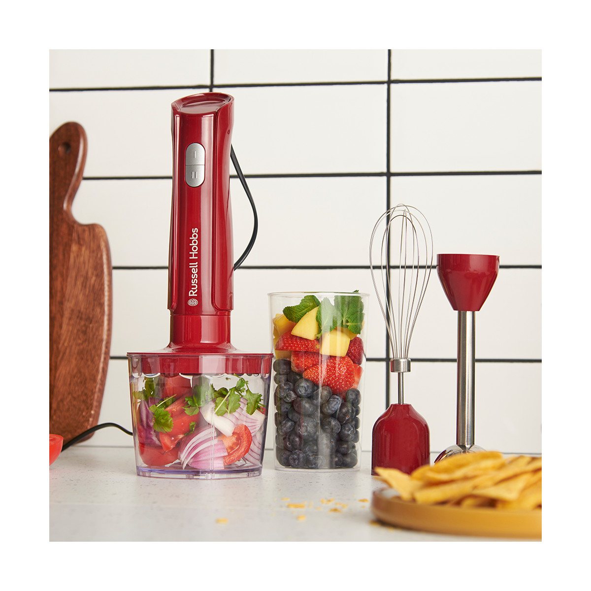 Ein roter Mixer mit Obst und Gemüse, ein Schneebesen und ein kleiner Foodprozessor stehen auf einer Theke. Holzschneidebrett im Hintergrund.