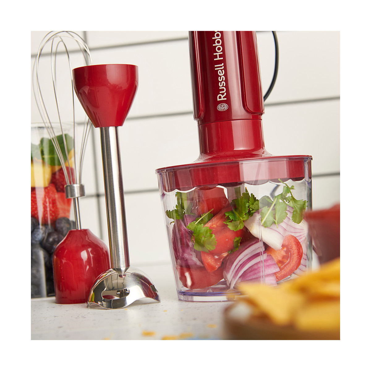 Ein roter Stabmixer mit einem Behälter mit gehacktem Gemüse und einem Glas mit Früchten und einem Schneebesen.