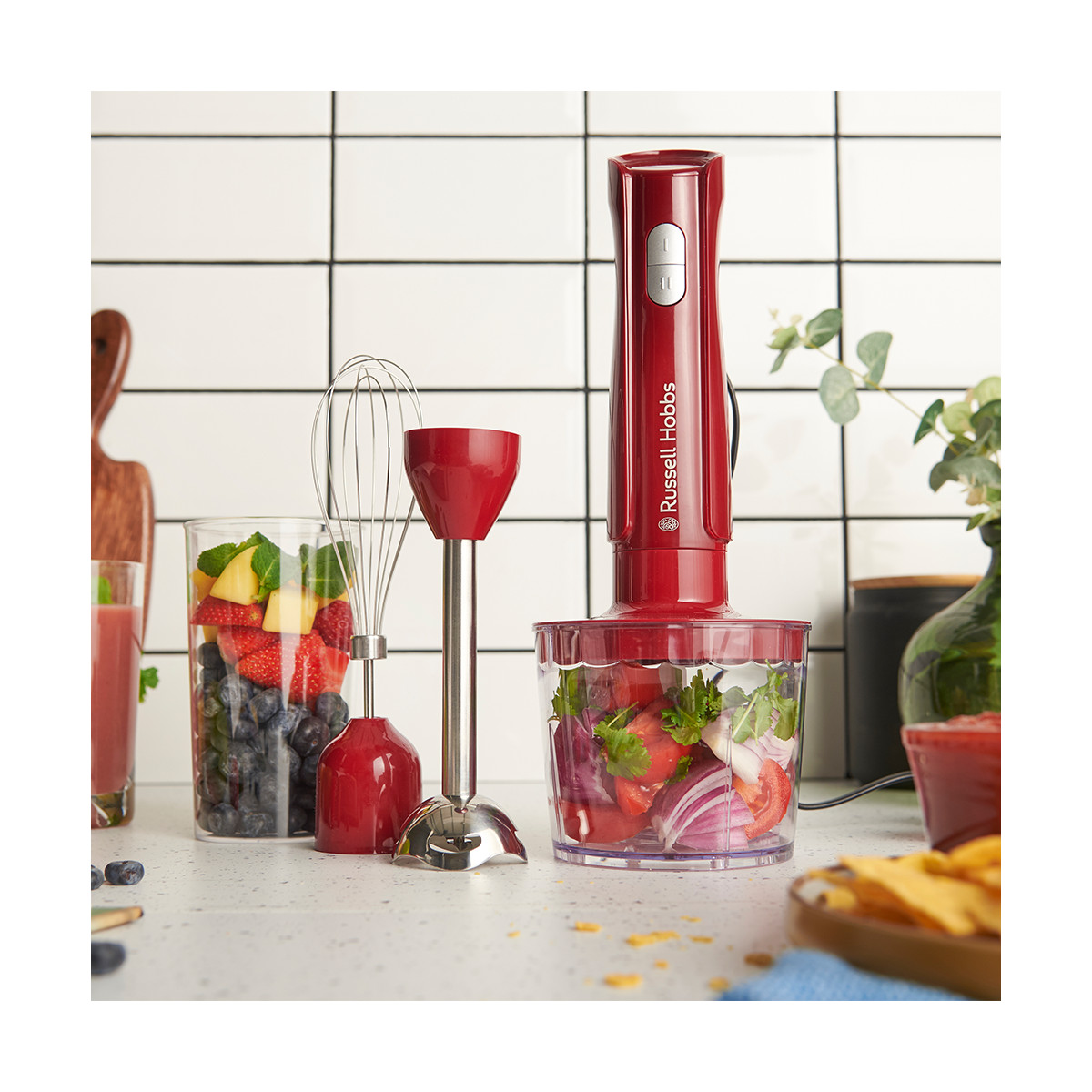 Ein roter Stabmixer mit Gemüse. Daneben ein Glas mit Früchten, ein Schneebesen und ein roter Behälter. Der Hintergrund hat Fliesen.