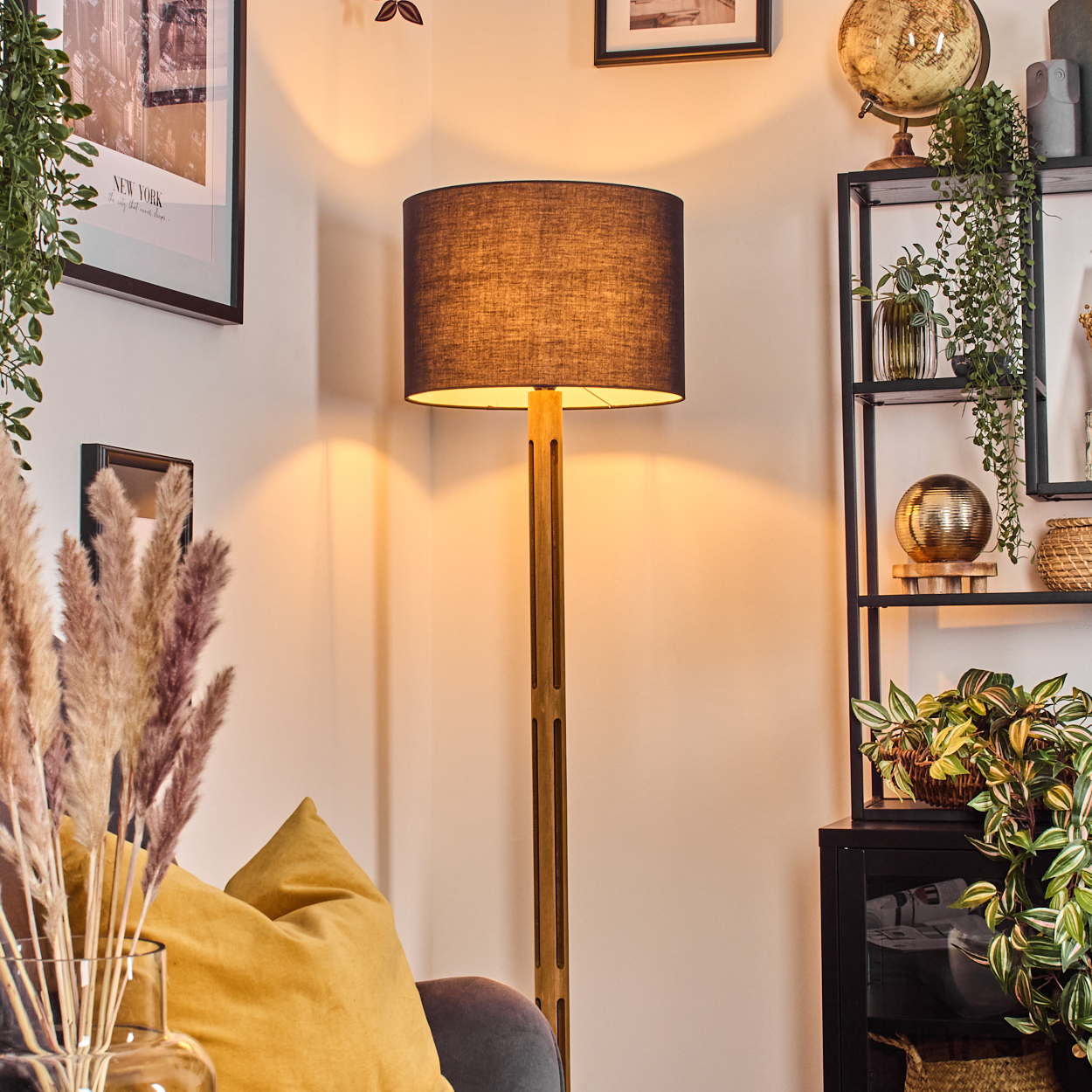Eine beleuchtete Stehlampe steht in einem Raum mit Regal, Pflanzen und Globus. Ein gelbes Kissen und gerahmte Kunst sind sichtbar.