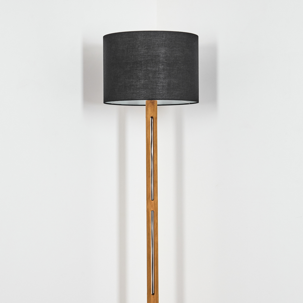 Eine Stehlampe mit schwarzem Lampenschirm und Holzständer steht vor einer weißen Wand. Die Lampe ist beleuchtet.
