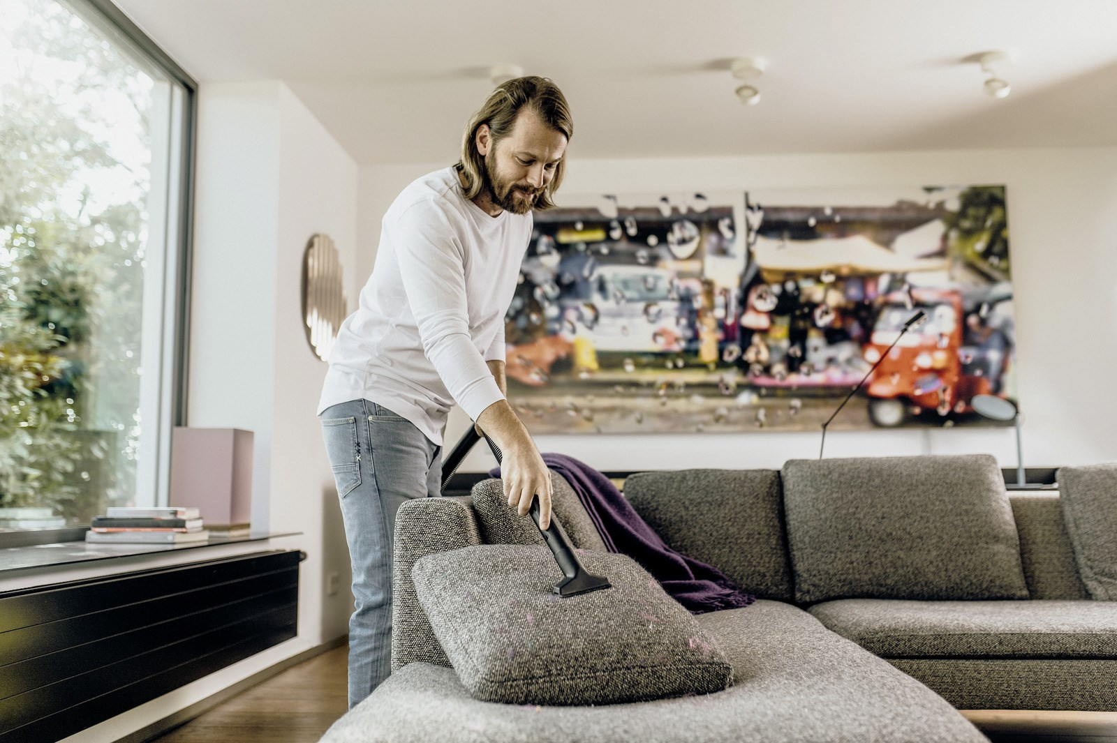 Mężczyzna odkurza poduszkę na kanapie. W pokoju jest duże okno, dzieła sztuki i fioletowy koc na kanapie.