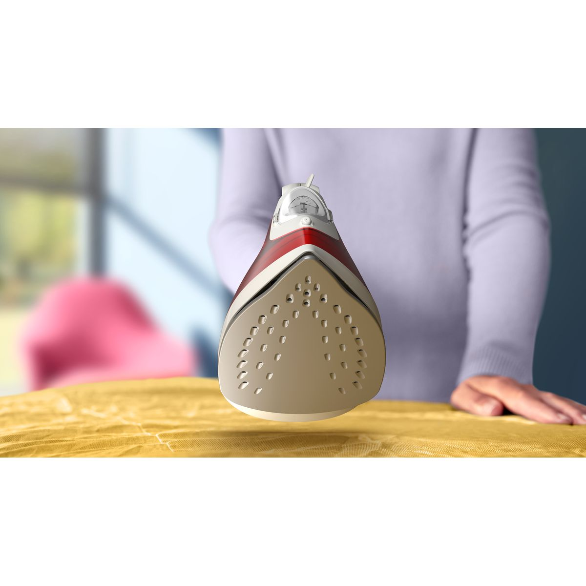 Zbliżenie białego i czerwonego żelazka unoszącego się nad żółtą deską do prasowania. Osoba w lawendowym swetrze w tle.