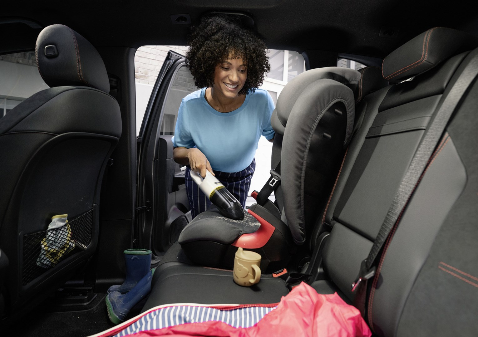 Kobieta odkurza wnętrze samochodu. Z tyłu fotelik dziecięcy. Są buty, kubek i czerwony koc.