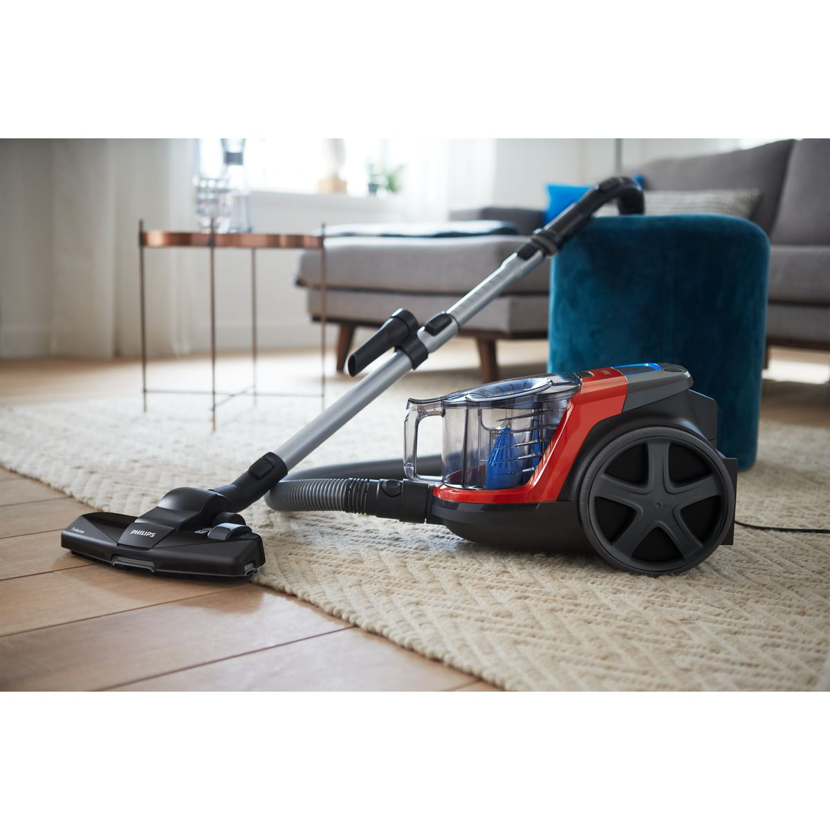Czerwony i czarny odkurzacz Philips stoi na beżowym dywanie. W tle sofa, stół i niebieska pufa.