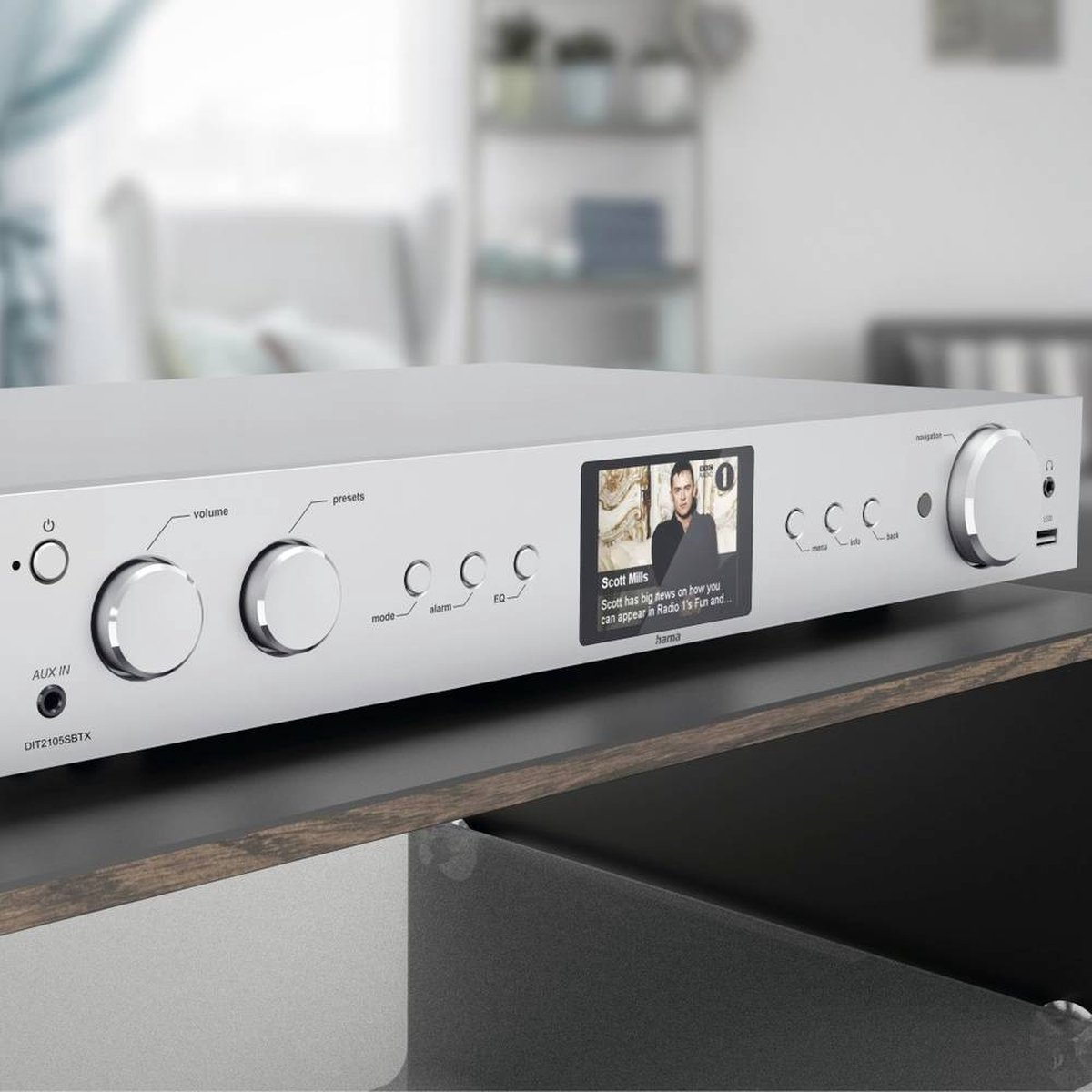 Srebrne urządzenie audio wyświetla mężczyznę na ekranie. Pokrętła i przyciski znajdują się z przodu. Umieszczone jest na drewnianej półce.