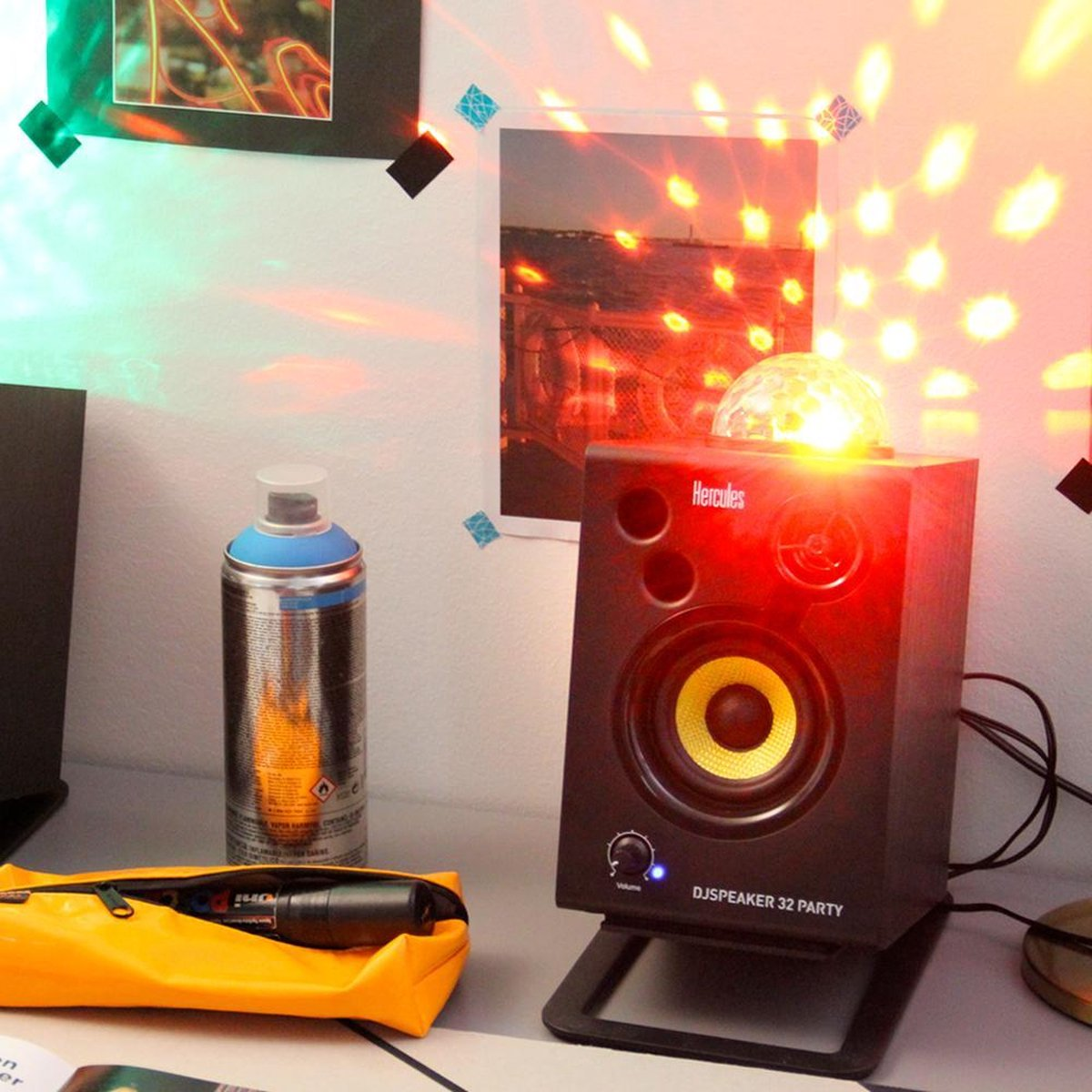 Een speaker met een discobal erop, spuitbus, gele etui en een foto aan de muur.