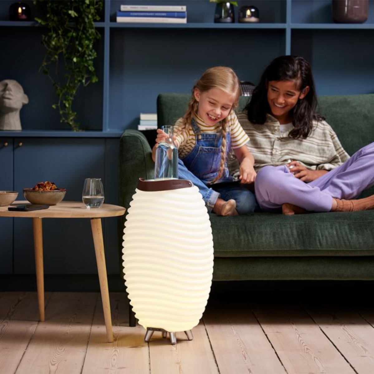 Twee meisjes zitten op een groene bank, een giet water in een lamp. Een bijzettafel met snacks en een glas is zichtbaar.