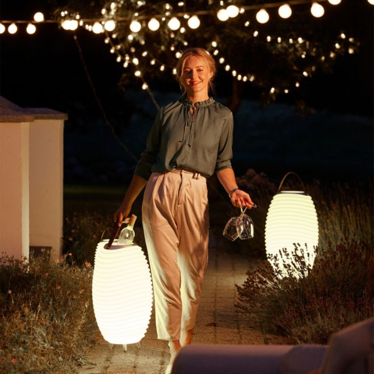 Een lachende vrouw houdt wijnglazen en lantaarns vast. Slingers met lichtjes erboven. Ze draagt een groen overhemd en een beige broek. Avondscène.