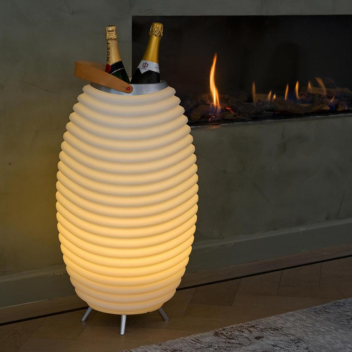 Een gloeiende, geribbelde lamp bevat twee champagneflessen. Een open haard met vuur staat op de achtergrond. De scène is binnen.