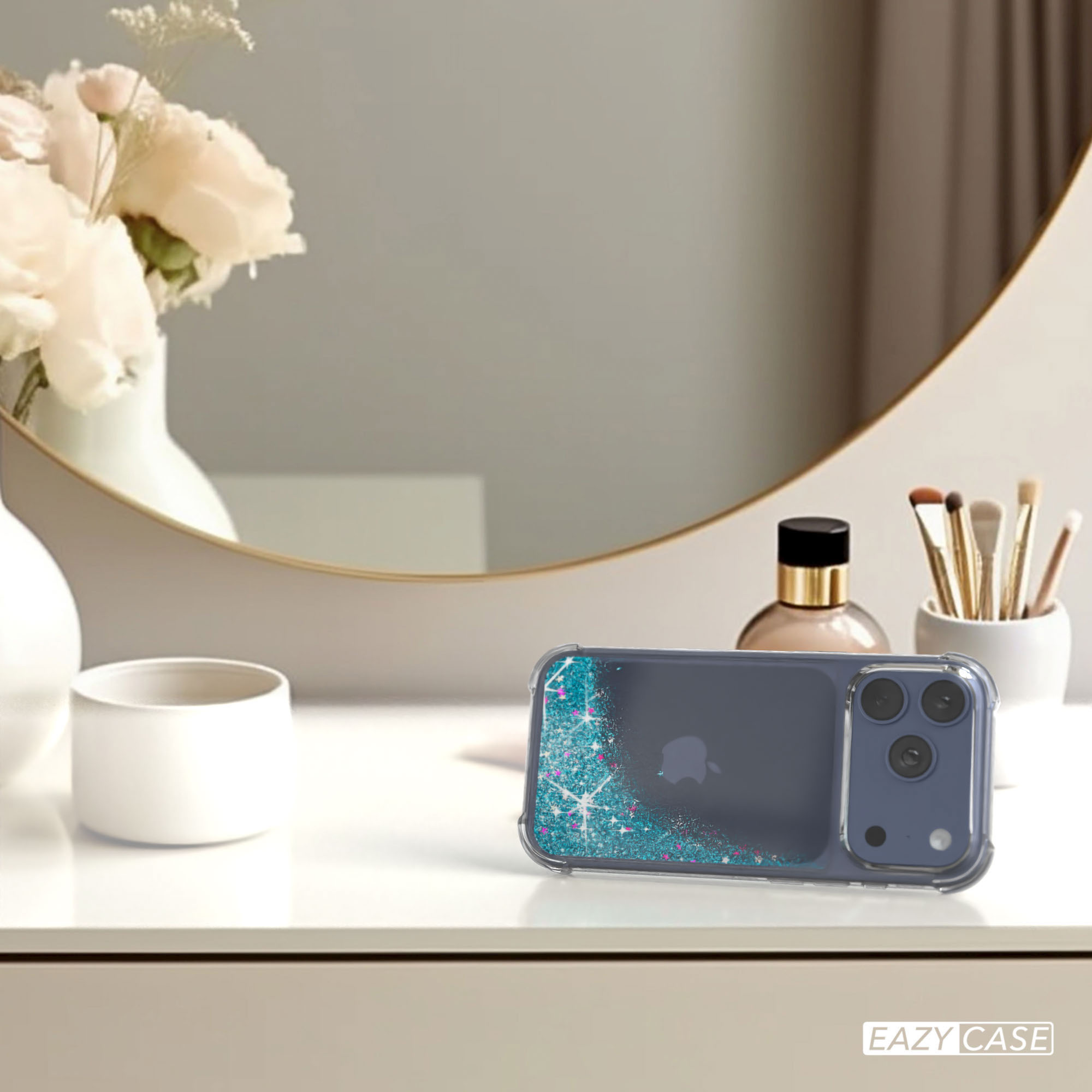 Ein iPhone in einer Glitzerhülle liegt auf einer weißen Oberfläche. Blumen, Spiegel, Parfüm und Pinsel im Hintergrund.